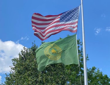 An American flag and JCC flag blowing in the wind together on a flag pole with a tree and blue sky in the background.