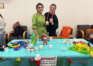 Two smiling students stand behind a table full of cups, Hawaiian leis, soda, heavy cream, and simple syrups. 