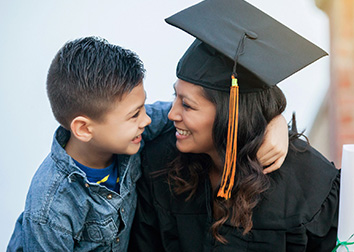 A young boy proudly looks at his mom as she is wearing a graduation cap and gown and holding a diploma.