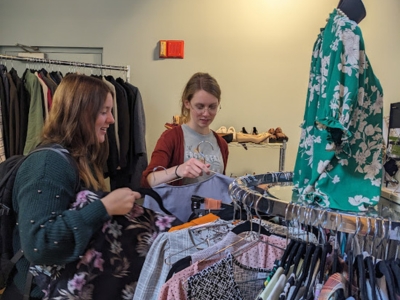 Two people hold clothing on hangars near a circular rack of clothing in a store-like room.