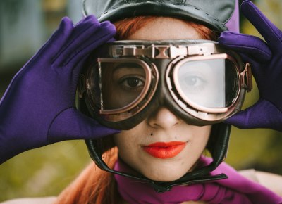 A person dressed in cosplay garb wearing large goggles and holding them with her gloved hands.