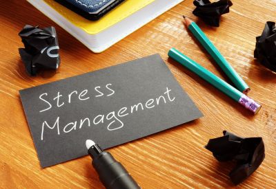 A black index card that reads stress management in white sitting a desk next to books, crumpled paper and a broken pencil.