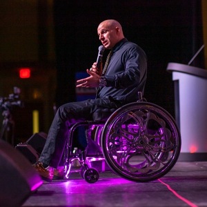 An image of Nick Scott on stage in his wheelchair with a purple light shining on him. He is wearing a gray suit, holding a microphone, and addressing an audience. 