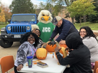Four students sit carving pumpkins at a table outdoors, with Eric Jones wearing a baseball cap, standing at the end of the table helping with one of the pumpkins. JJ Jayhawk is to his right, wearing a green JCC t-shirt, and there is a blue jeep to JJ's right. 