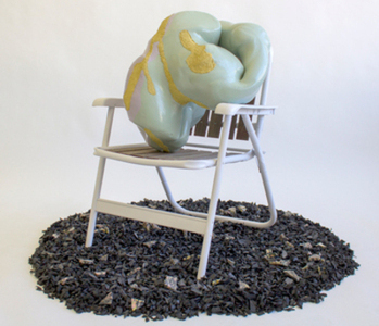 A sculpture positioned in a garden chair, situated on material that resembles dark brown mulch.