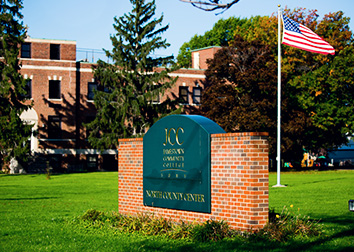 A view of the SUNY JCC sign at the North County Center that shows an American flag, pine trees, and the main building in the background.