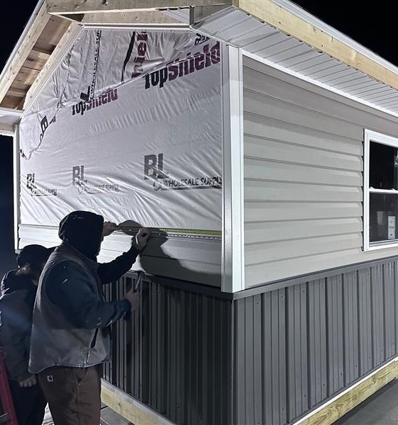 Two people, wearing hooded jackets, measure the siding on a shed building at night. The structure has partial vinyl siding, metal panels, and exposed insulation wrap.