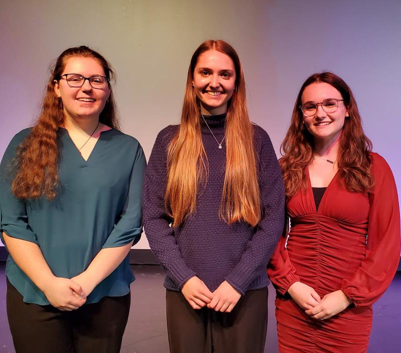 Three smiling students pose together for a photo on a stage.