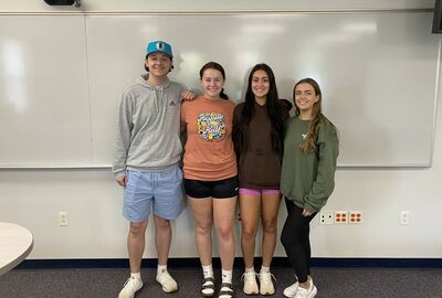 Four students pose with arms wrapped around each other in a classroom with a large white dry erase board in the background.