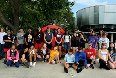 A large group of students poses together on a sidewalk outside the Corning Museum of Glass.